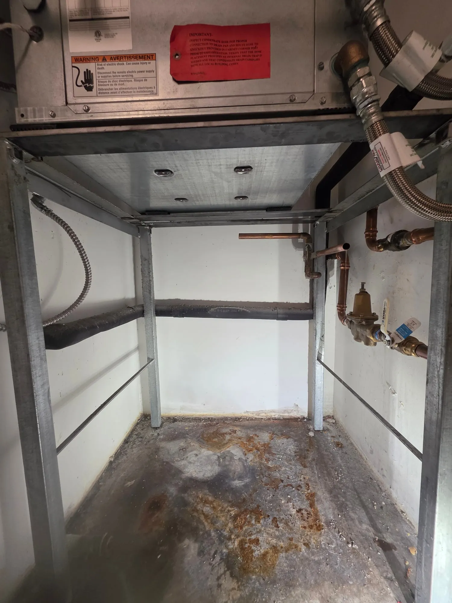 Empty water heater stand under an air handler with rust stains on the floor