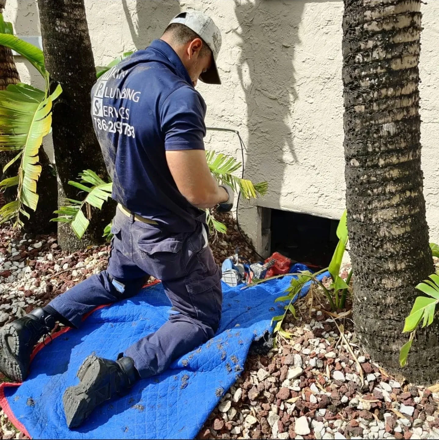 Plumber kneeling at a crawlspace access beside palm trees